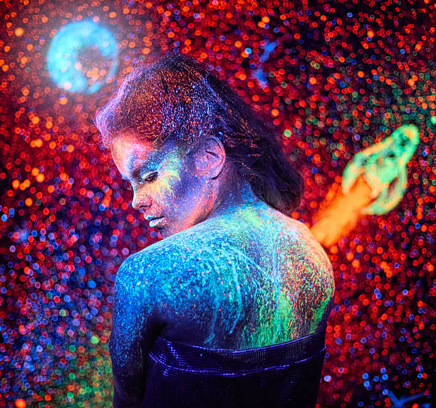 Beautiful young woman with neon fluorescent painting on a face (the makeup) and on a body (the body art) is standing on background with image luminous starry sky with rocket. Studio shooting in ultraviolet lighting