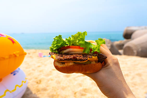 istock Woman holding burger in a hand against beach background 2230560397
