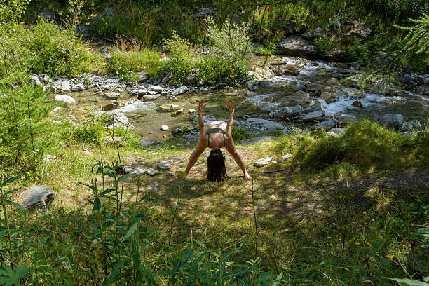 A woman practices a wide-legged forward fold yoga pose outdoors next to a clear, rocky mountain stream. The serene setting and the woman's focused expression create a sense of calm and mindfulness in nature, ideal for wellness and healthy lifestyle concepts.