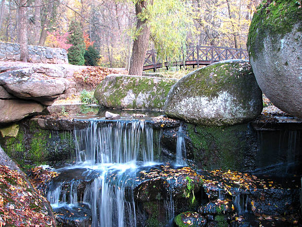 istock Small waterfall among mossy rocks in autumn forest park with stone bridge on background, symbol of tranquility, nature harmony, peaceful landscape and scenic seasonal beauty of nature 2230567042