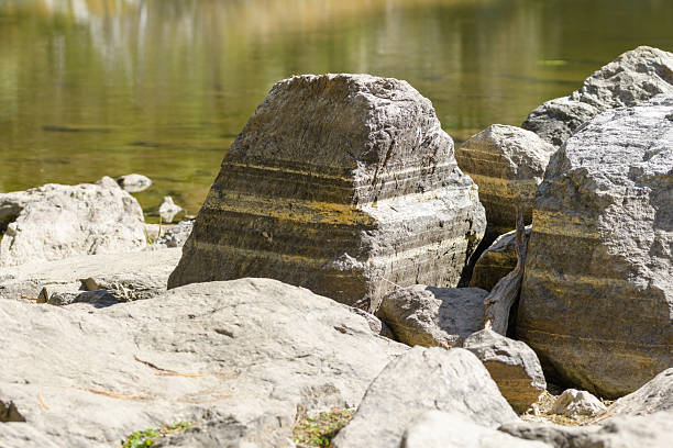 A close-up photograph showcases a collection of river rocks along a shoreline, with distinct geological layers visible in the primary rock. The background features blurred water, enhancing the natural and serene landscape, ideal for environmental or geology-themed projects.