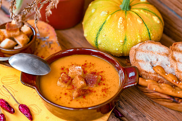 Rustic autumn table setting with a bowl of creamy pumpkin soup topped with golden croutons and paprika. Served with toasted bread and breadsticks, surrounded by fresh pumpkin and cozy seasonal decor, this warm dish represents comfort food, fall flavors, and homemade cooking.
