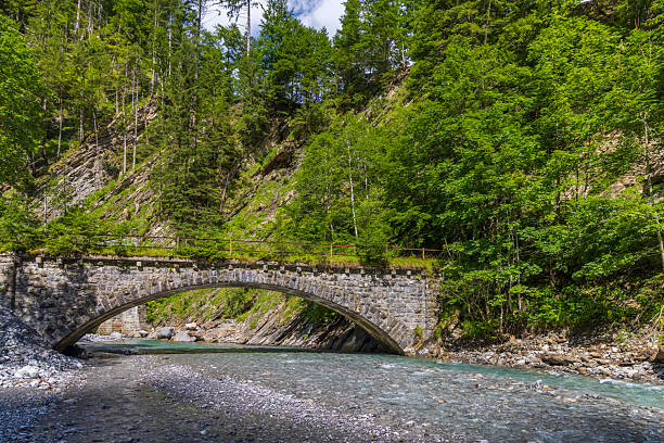 Scenic view of an old stone bridge over a clear river flowing through a lush green forest in Bregenzerwald, Vorarlberg, Austria