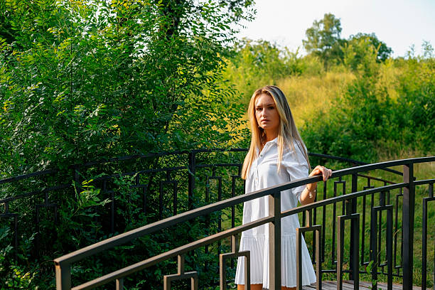 Portrait of a beautiful young blonde woman standing on a bridge in a park during a sunny summer day