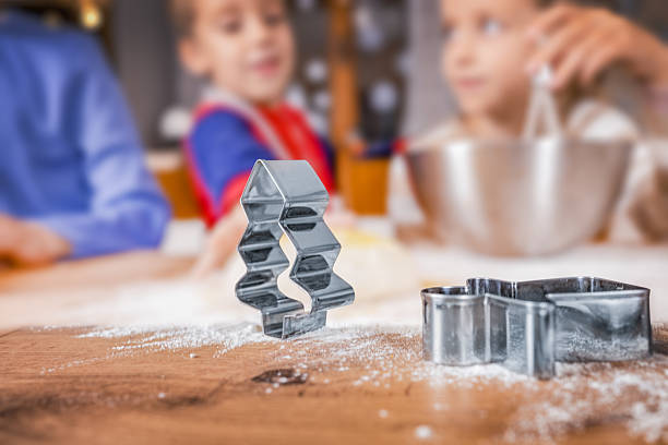 Close-up of a Christmas tree-shaped cookie cutter on a wooden table sprinkled with flour, with blurred children in the background preparing dough for holiday cookies. Festive family baking activity symbolizing Christmas traditions, childhood joy, and holiday spirit.