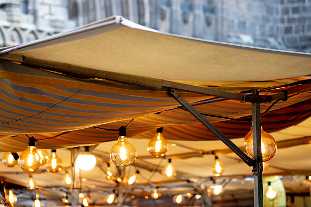 Atmospheric view of glowing string lights hanging under a striped canopy at an outdoor market in Barcelona, Spain. The warm bulbs create a festive and cozy ambiance, reflecting the charm of Mediterranean evenings, local gatherings, and street culture.