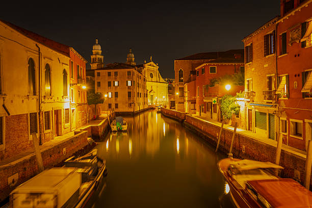 Quiet Venetian canal at night with moored boats, historic houses, and a small stone bridge in the distance.