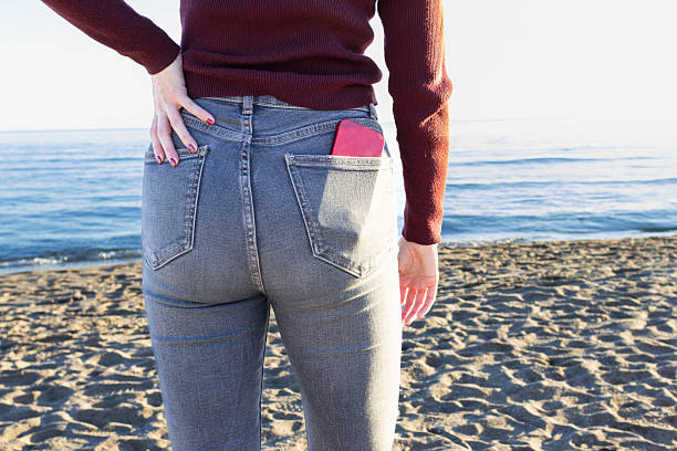 Woman wearing jeans, with a vibrant red smartphone tucked in her back pocket, standing on the beach and gazing thoughtfully at the serene sea under a clear blue sky
