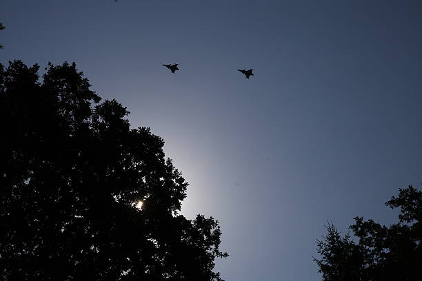 Two Saab JAS 39 Gripen fighter jets are captured in flight above a forest canopy during a clear sunny day. The sun shines through the trees, casting dramatic rays and emphasizing the contrast between the stillness of nature and the speed of the jets. A visually compelling mix of military power and natural beauty.