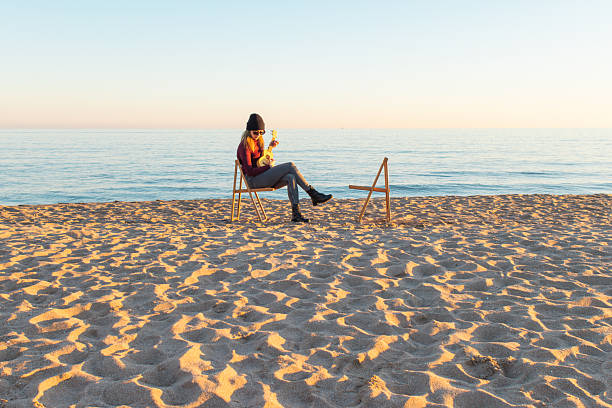 adult woman sitting on a wooden chair playing ukulele on a sandy beach at sunset, with a empty wooden chair next to her, enjoying mindfulness.