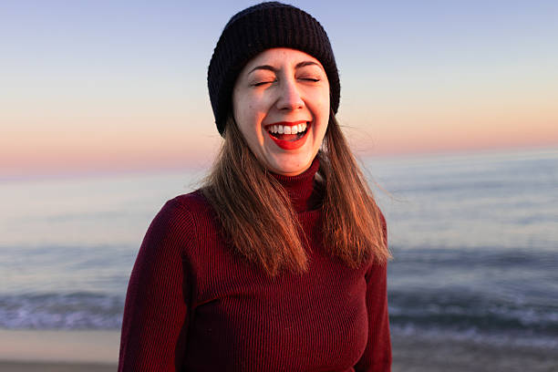 Young woman laughing with closed eyes on the beach during a stunning sunset, embracing feelings of freedom and joy while wearing stylish winter clothes and red lipstick