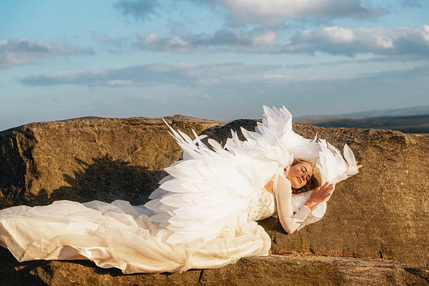 A woman dressed in an elegant white gown and intricately designed feathered wings lies peacefully on a rock against a picturesque backdrop of clouds and hills.