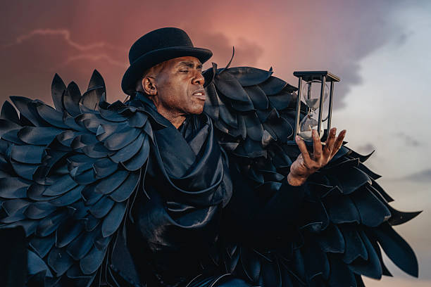 A performer in a black feathered costume holds a lantern against a dramatic sunset sky during a theatrical event in an outdoor setting
