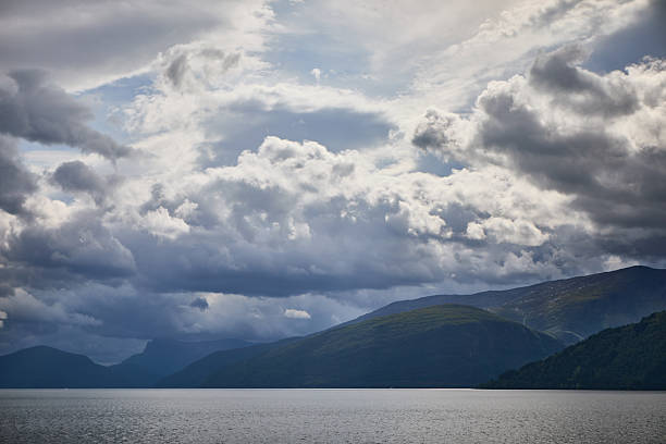 A breathtaking view of a serene lake with dramatic clouds and mountainous terrains under a moody sky, evoking tranquility and awe at nature's beauty.