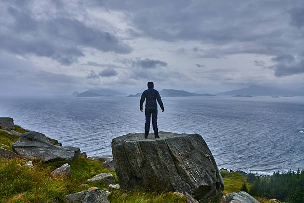 A solitary hiker stands atop a large rock, gazing at a scenic coastal view under a cloudy sky. The image captures solitude, exploration, and the natural beauty of rugged landscapes along the water.