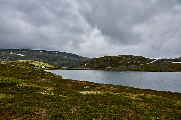 A tranquil mountain lake surrounded by rugged terrain, under a brooding overcast sky, highlights a desolate yet serene landscape. A distant road curves through the wilderness, adding context to the scene.