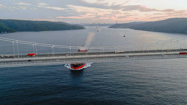 Aerial view of a large bulk carrier cargo ship with a prominent helipad navigating beneath the Yavuz Sultan Selim Bridge over the Bosphorus Strait in Istanbul, Turkey. The red vessel, designed for transporting solid bulk goods, sails at sunset while road traffic crosses the suspension bridge overhead. The dramatic juxtaposition of maritime and roadway infrastructure showcases global logistics, international trade, and seamless connectivity between continents.