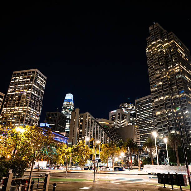 Nighttime view of downtown San Francisco with illuminated skyscrapers, palm trees, and light trails from moving cars. The urban cityscape highlights the vibrant energy and architecture of the financial district.