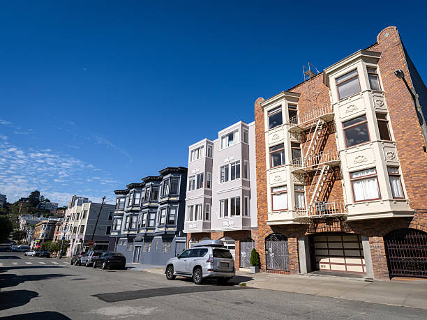 A residential building with a traditional fire escape ladder stands under a clear blue sky in San Francisco. The street is lined with parked cars and colorful facades, capturing the charm of the city's distinctive architecture.