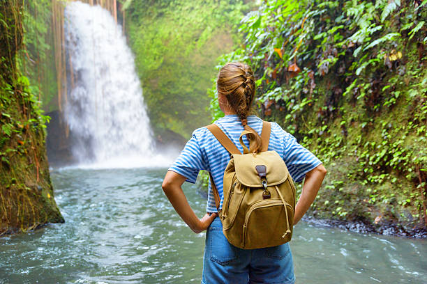 Female tourist with backpack enjoying view of awesome waterfall. Woman standing in water of river among tropical forest. Fairy landscape. Amazing Asian trip. Solo traveler. Freedom concept.