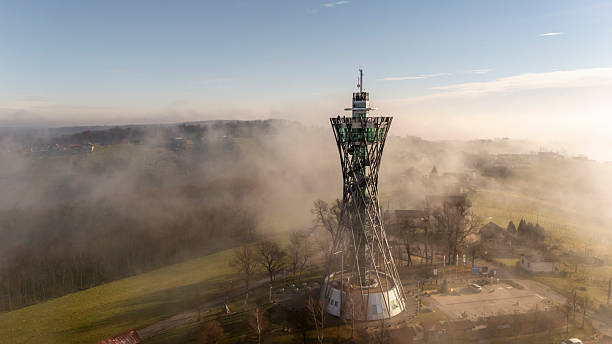 A communication tower is standing tall amidst a foggy landscape at sunrise. The surrounding area is enveloped in mist, creating a serene and atmospheric setting with scattered trees and open fields.