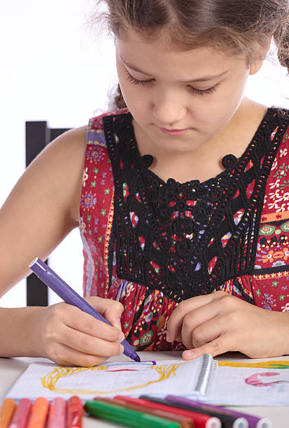 Cute elementary age girl with two funny pigtails is sitting at a table and drawing with colored markers in notebook. Studio shooting on white background