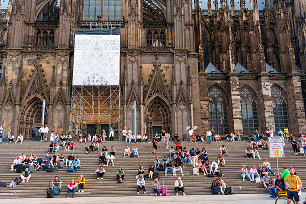 Cologne, Germany - June 15, 2025: Vibrant scene of visitors sitting on steps of Cologne Cathedral, showcasing Gothic architecture with intricate details and bustling city life.