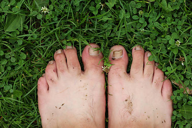 Bare feet with a few dark dirt spots rest on dense green grass mixed with white clover. The image conveys a sense of freedom, connection with nature, and simple, everyday relaxation.
