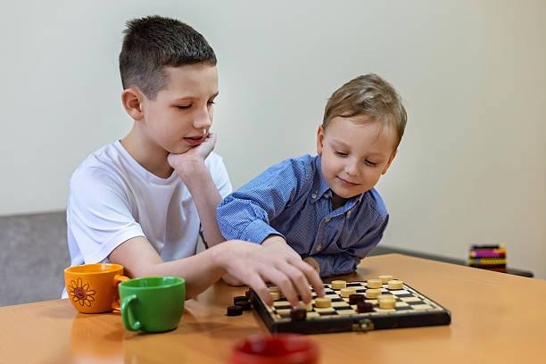 Two Boys Playing Checkers at Home. Two young boys playing a game of checkers at a table, focused and engaged, surrounded by toy cars and colorful mugs.