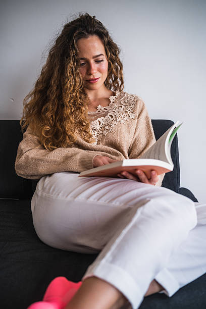 istock Young woman reading a book on the sofa at home 2230724406