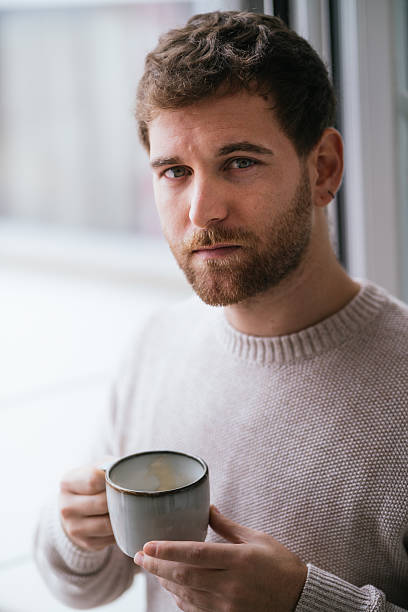 istock Young man drinking coffee by window in cozy sweater 2230726037