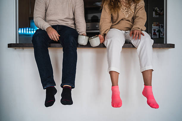 Young couple sitting on kitchen counter dangling their feet, wearing colorful socks and holding coffee mugs, enjoying a relaxing moment together