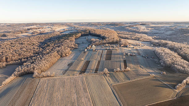A frosty landscape is stretching across fields and forests at sunrise. The fields are covered with a light frost, and the trees are appearing bare in the early morning light. Small buildings are scattered throughout the rural area.