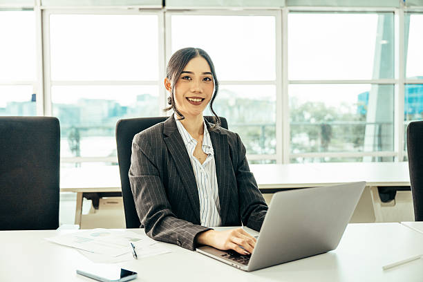 istock Asian businesswoman smiling while at work in a modern office with a laptop and documents on the desk 2230735100