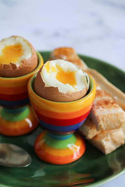 Close-up image of rainbow colour striped egg cups with soft boiled eggs, white buttered toasted bread soldiers on green tea plate, stainless steel teaspoon, white marble effect background, elevated view, focus on foreground