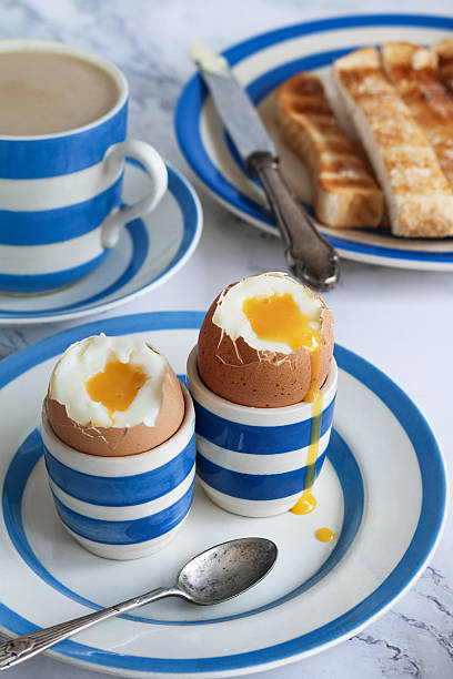 Stock photo showing close-up view of a breakfast scene on a white marble effect surface. The scene is set with Cornish ware crockery of tea plates containing egg cups of soft boiled eggs and white buttered toasted bread soldiers along side hot, milky tea in a blue and white striped teacup and saucer.