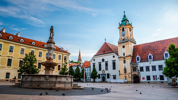 Main Square (Hlavné námestie) in Bratislava’s Old Town, featuring the Maximilian Fountain in the foreground and the historic Old Town Hall with its iconic clock tower in the background. The square is surrounded by colorful historic buildings, captured in soft morning light. Bratislava, capital city of Slovakia in Europe.