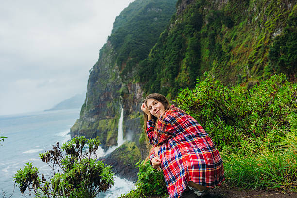 Portrait of handsome female with long hair wearing red shirt admiring majestic views of green mountains and waterfall falling into the ocean on Madeira island, Portugal