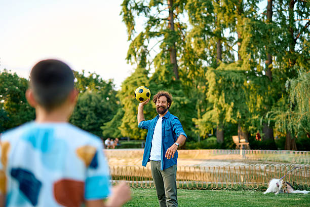 Father and son enjoying a sunny day in the park, playing soccer and creating joyful memories while strengthening their bond amidst the vibrant green grass and trees
