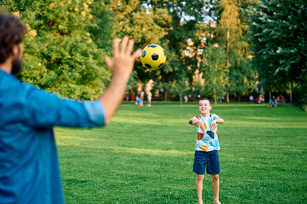 Father and son enjoying a sunny summer day in the park, playing together with a soccer ball, sharing moments of joy and bonding amidst the green grass and trees