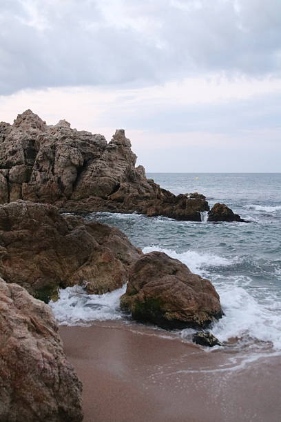 View from the shore of clear blue waves flowing between rocks, with a bright cloudy sky on the horizon. The serene scene captures the dynamic interaction between sea and coastline.