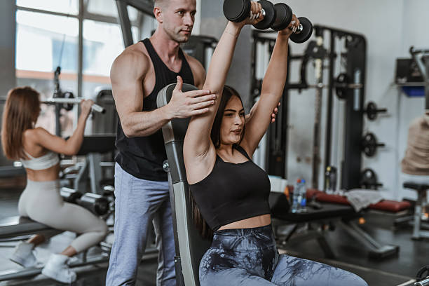 istock Trainer Helping Woman Execute Shoulder Press With Dumbbells While Standing Behind Her At Gym 2230770891