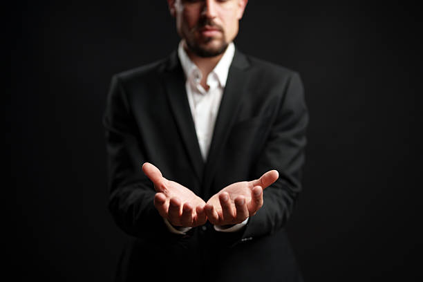 A businessman stands confidently with his hands open toward the viewer, dressed in a tailored suit and shirt. The dark background emphasizes his professional demeanor and focus.