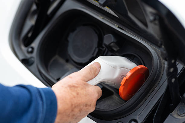 A close-up shot of a man plugging in a charging cable to a white electric car outside in a public car park. The car is being charged using a public electric charging port.Videos are available similar to this scenario.