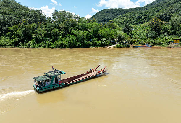 A vibrant and authentic scene of a small ferry boat navigating the wide, muddy waters of the Mekong River. The boat, with its distinctively utilitarian design, is positioned in the center of the frame, transporting people or goods between the riverbanks. The background features a lush, green, and forested hillside under a clear blue sky, characteristic of the Southeast Asian landscape. The foreground shows the riverbank with some low-lying vegetation, adding depth to the image. This picture captures a slice of daily life and transport on one of the world's great rivers.