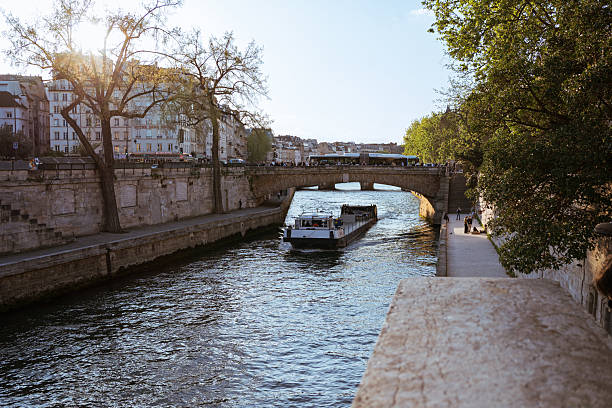 Paris, France - April 17, 2025: Seine river scene with sightseeing boat passing under Pont Marie. People walk along the riverside promenade. Classic Parisian buildings and trees at golden hour.
