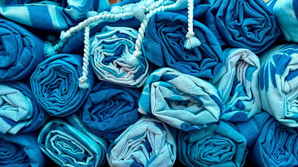 This is a close-up, top-down shot of several tightly rolled towels or blankets in various shades of blue. The image is filled with the circular, spiral patterns created by the rolls, which are packed together, creating a sense of texture and depth. The color palette is composed of different tones, from deep navy and royal blue to lighter shades of sky blue and almost white. The fabric has a visible woven texture, adding to the visual interest. The composition is an abstract and calming pattern, evoking feelings of comfort, relaxation, and cleanliness, making it an excellent image for themes related to home decor, spas, travel, or laundry.