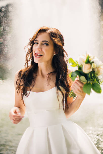A happy bride in a white gown smiles over her shoulder holding a bouquet. Bright, backlit scene with a fountain creating a dreamy, romantic mood. Vertical portrait with shallow depth of field