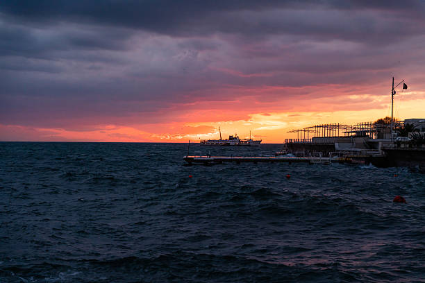 A scenic view of a ferry sailing on the Bosphorus during sunset in Istanbul, Turkey. The dramatic sky with orange and purple tones, the rough sea, and the silhouette of the pier create a timeless seascape.
