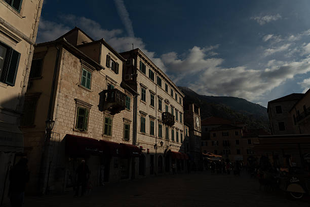 Scenic view of Kotor Old Town square at twilight, featuring the historic clock tower, stone houses with green shutters, and the surrounding mountains. The warm evening light and shadows highlight the charm of this UNESCO World Heritage site in Montenegro.